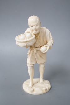 A Japanese carved ivory figure, Meiji period, modelled as a fruit vendor carrying a bowl of fruit in one hand and a piece of fruit in the other, standing on a naturalistic circular base, signed on base, height 14.5cm